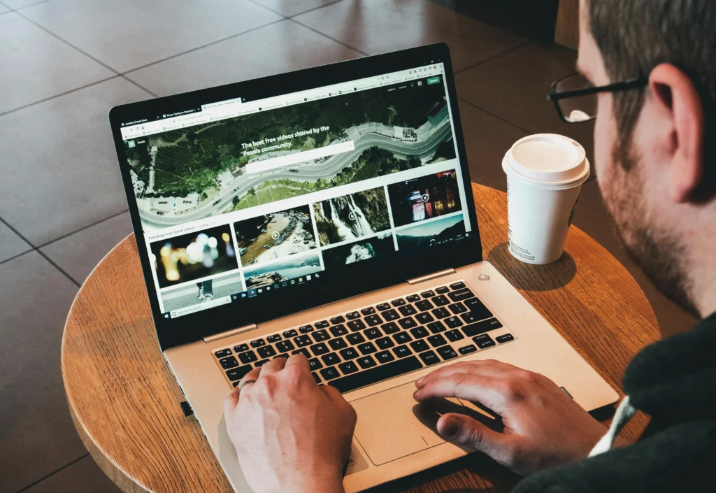 A man working on a laptop in a café, browsing a website with images and videos, with a cup of coffee beside him.