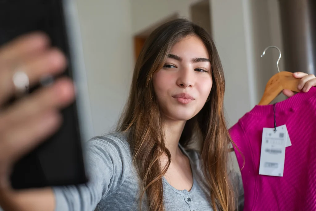 Young woman holding a pink top on a hanger while looking at her phone, possibly reviewing or promoting the product online.